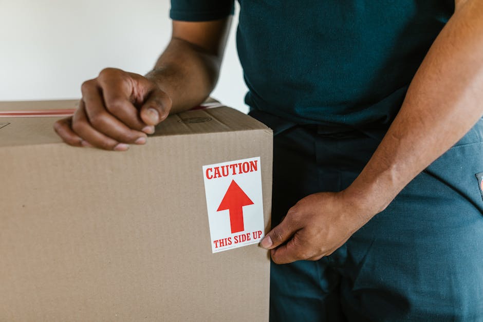 Close-up image of a cardboard moving box labeled with handwritten text in blue ink that reads 'KITCHEN' indicating the contents are kitchen items. The box also has printed sections for 'NAME,' 'CONTEN